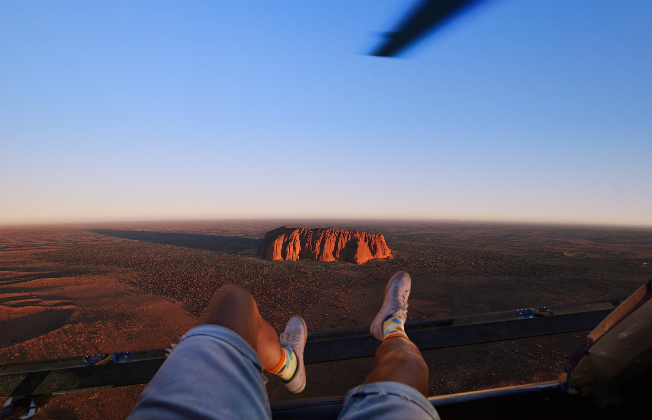 Point of view of person looking down at Uluru from a helicopter. Person's legs are hanging over